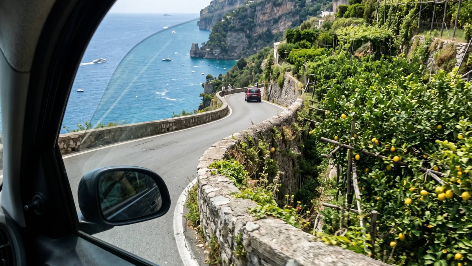A winding coast road above a blue sea, seen through a car window, with lemon trees on the terraces beside it